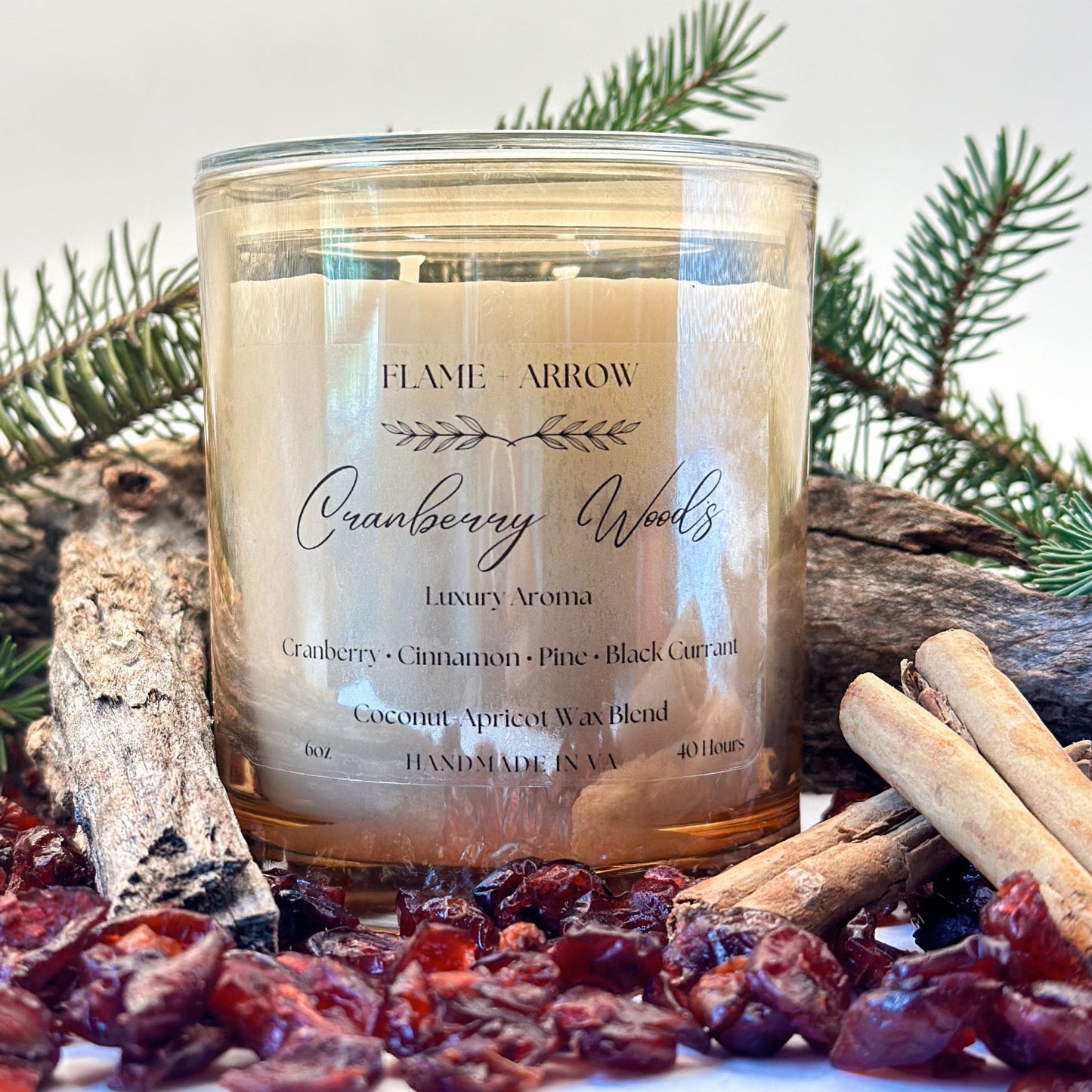 A champagne glass jar candle with a label reading 'Cranberry Woods Luxury Aroma'. It is surrounded by cranberries, pine branches, and a cinnamon stick, suggesting a cranberry, cinnamon, pine, and black currant scent.
