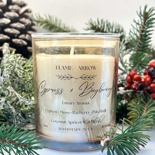 A champagne jar candle with 'Cypress + Bayberry' labeled on it, surrounded by pine branches and decorative pine cones.