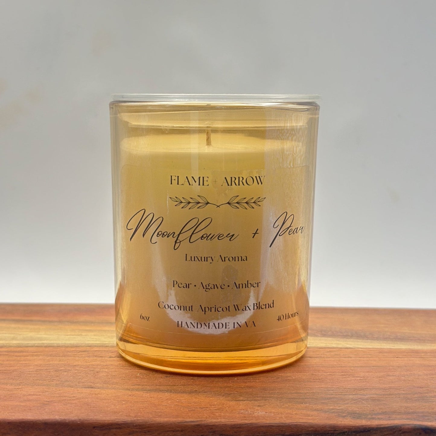 A clear glass candle jar with a label that reads 'Flame Arrow Moonflower Pear Luxury Aroma', with additional scent notes listed below.