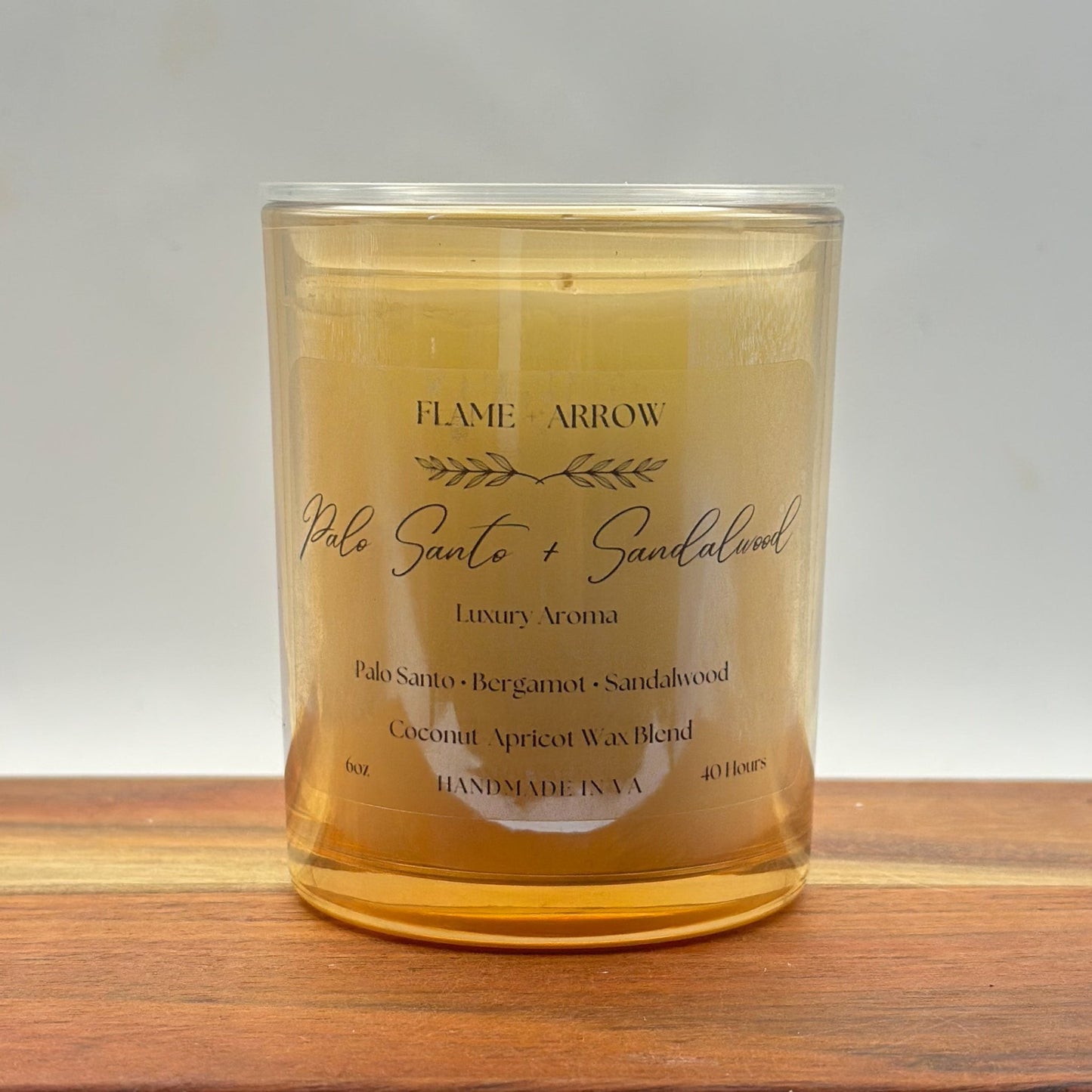 A clear glass candle jar with a label that reads 'Flame Arrow Palo Santo 
and Sandalwood Luxury Aroma', with additional scent notes listed below.