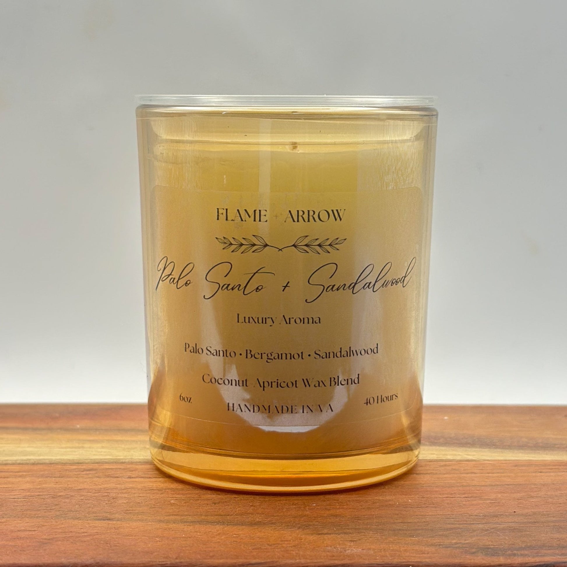 A clear glass candle jar with a label that reads 'Flame Arrow Palo Santo 
and Sandalwood Luxury Aroma', with additional scent notes listed below.