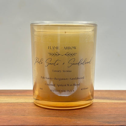 A clear glass candle jar with a label that reads 'Flame Arrow Palo Santo 
and Sandalwood Luxury Aroma', with additional scent notes listed below.